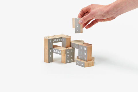 A hand is seen placing a gray block onto a structure made from brown and gray wood building blocks designed to represent architectural brutalism elements. 