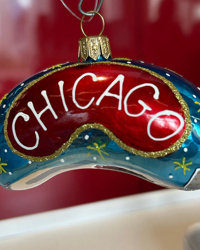 A hand-painted glass Chicago-themed ornament shaped like a curved game piece, featuring a red center with white 'CHICAGO' lettering, a blue outer section with gold stars, and a silver-and-blue cityscape detail, hanging against a red and beige background.