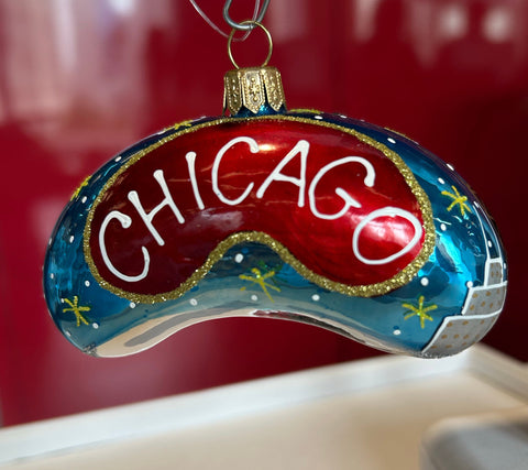 A hand-painted glass Chicago-themed ornament shaped like a curved game piece, featuring a red center with white 'CHICAGO' lettering, a blue outer section with gold stars, and a silver-and-blue cityscape detail, hanging against a red and beige background.