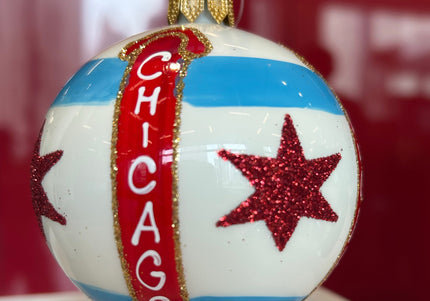 A spherical Christmas ornament featuring the Chicago flag design, with a vertical red banner reading 'CHICAGO' and two red glitter stars, hanging against a red background.