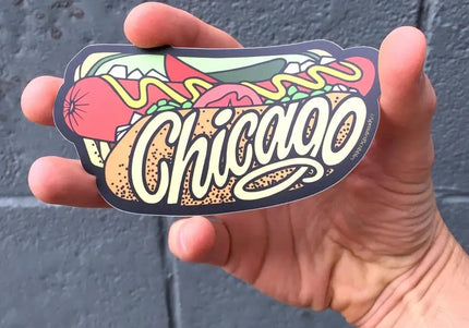 Hand holding a hot dog 'Chicago' sticker against a gray wall.