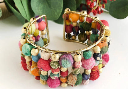 A colorful beaded cuff bracelet made from layers of Kantha beads with gold accents, displayed against a white background with green foliage in the background.