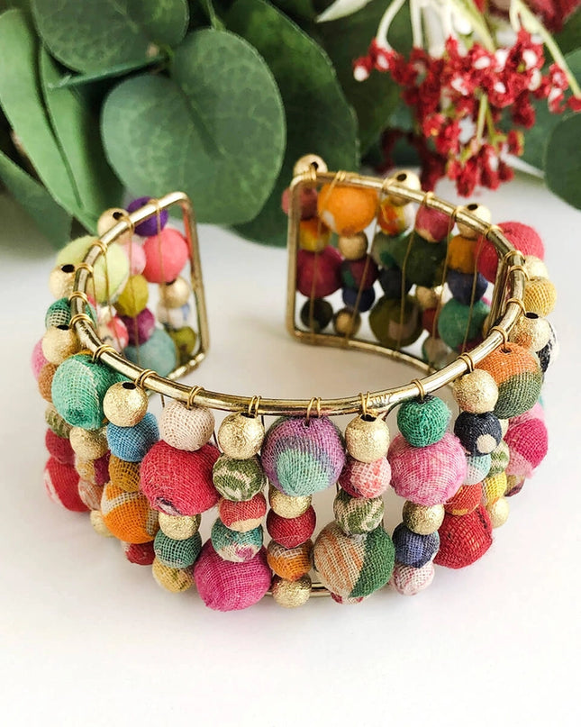 A colorful beaded cuff bracelet made from layers of Kantha beads with gold accents, displayed against a white background with green foliage in the background.
