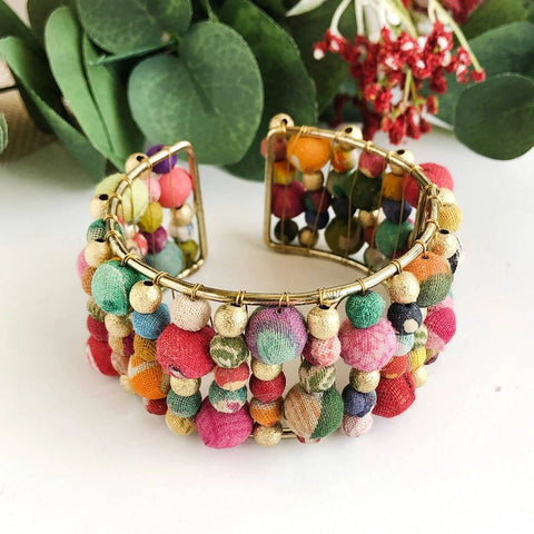 A colorful beaded cuff bracelet made from layers of Kantha beads with gold accents, displayed against a white background with green foliage in the background.